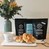 Hjertekopp, kanelboller og tre pakker med Røverkaffe såtr på et bord med blomster